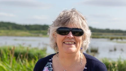 Blind veteran Christine, wearing dark sunglasses and holding a guide cane, as she smiles for a photograph in front of tall grass and a lake in the background