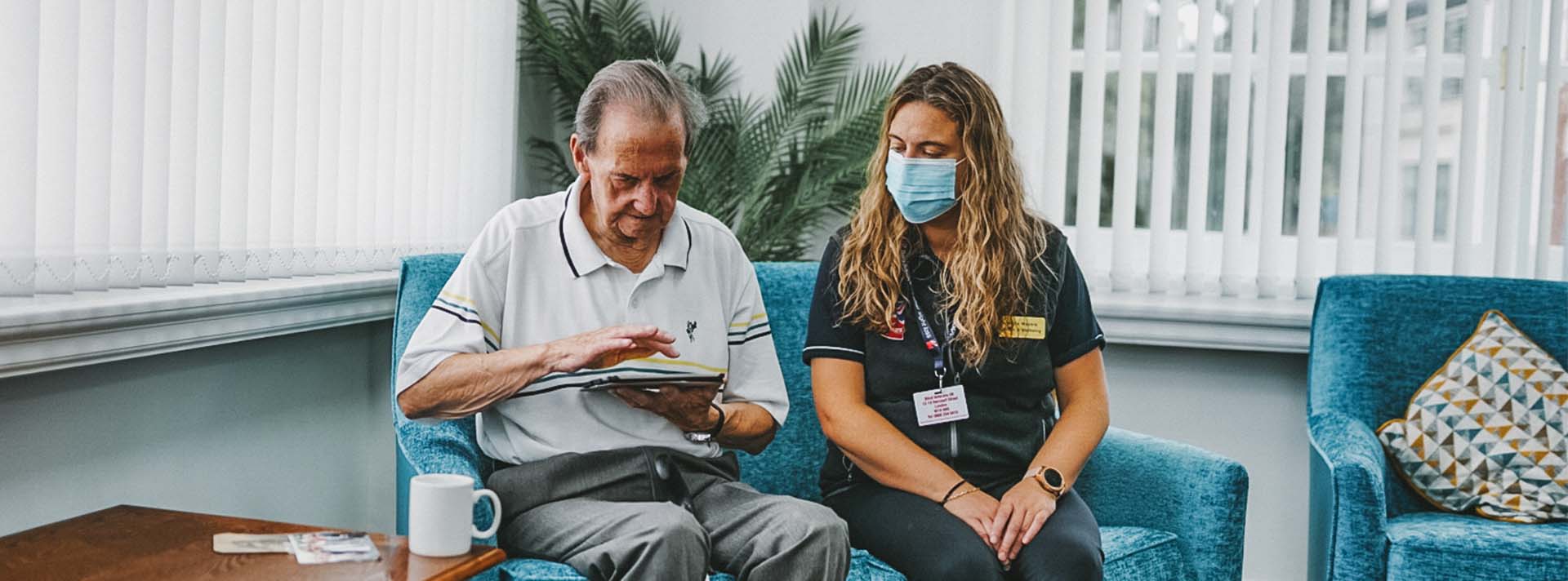 Blind veteran veteran Fred using a tablet, while sitting on a sofa with a Blind Veterans UK staff member