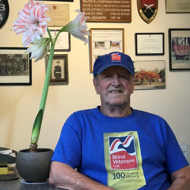 John is sitting next to a blooming amaryllis, as he wears a Blind Veterans UK blue top. Behind him, the wall is adorned with various military memorabilia.