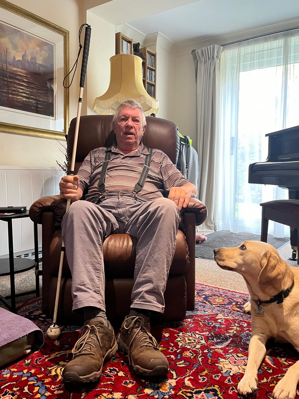 Graham sitting at home holding his white cane with his guide dog by his side