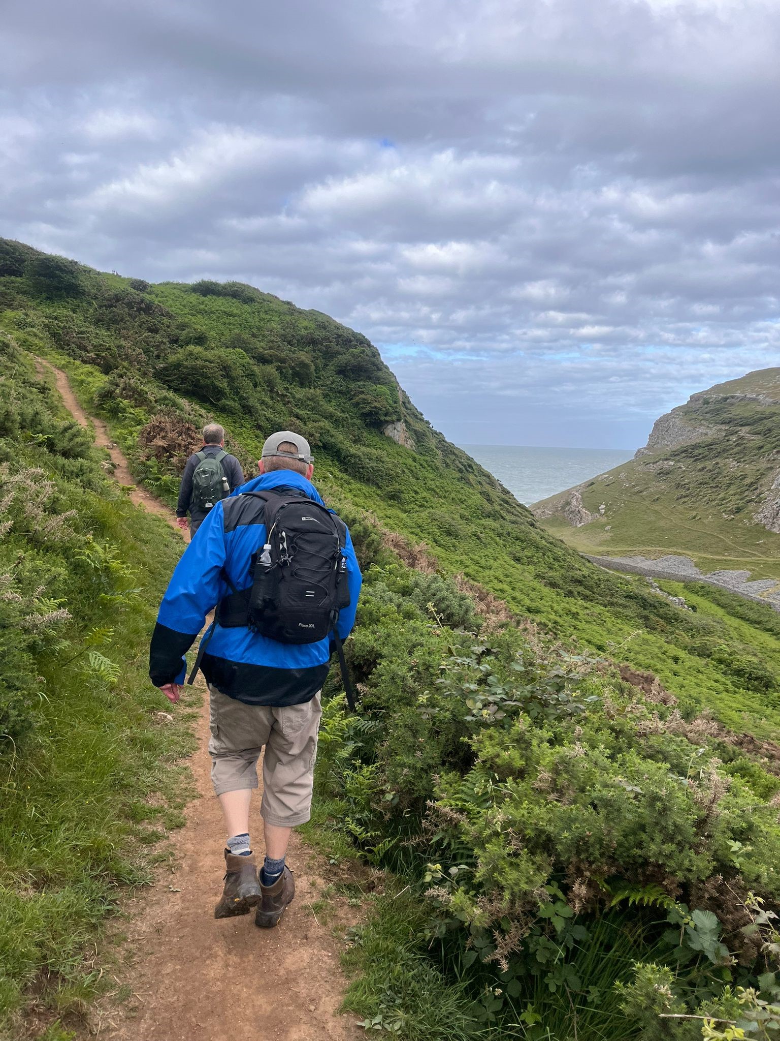 A picture of Phil from behind as he uses his white cane to navigate along a thin pathway which is going uphill along the coastline