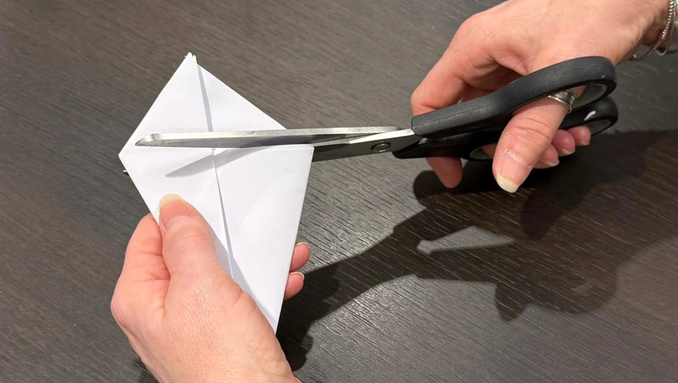 The image shows a person holding a folded triangle of paper in one hand and using scissors in the other hand to cut off the pointed ends. This step prepares the paper for the next stage of creating a snowflake.