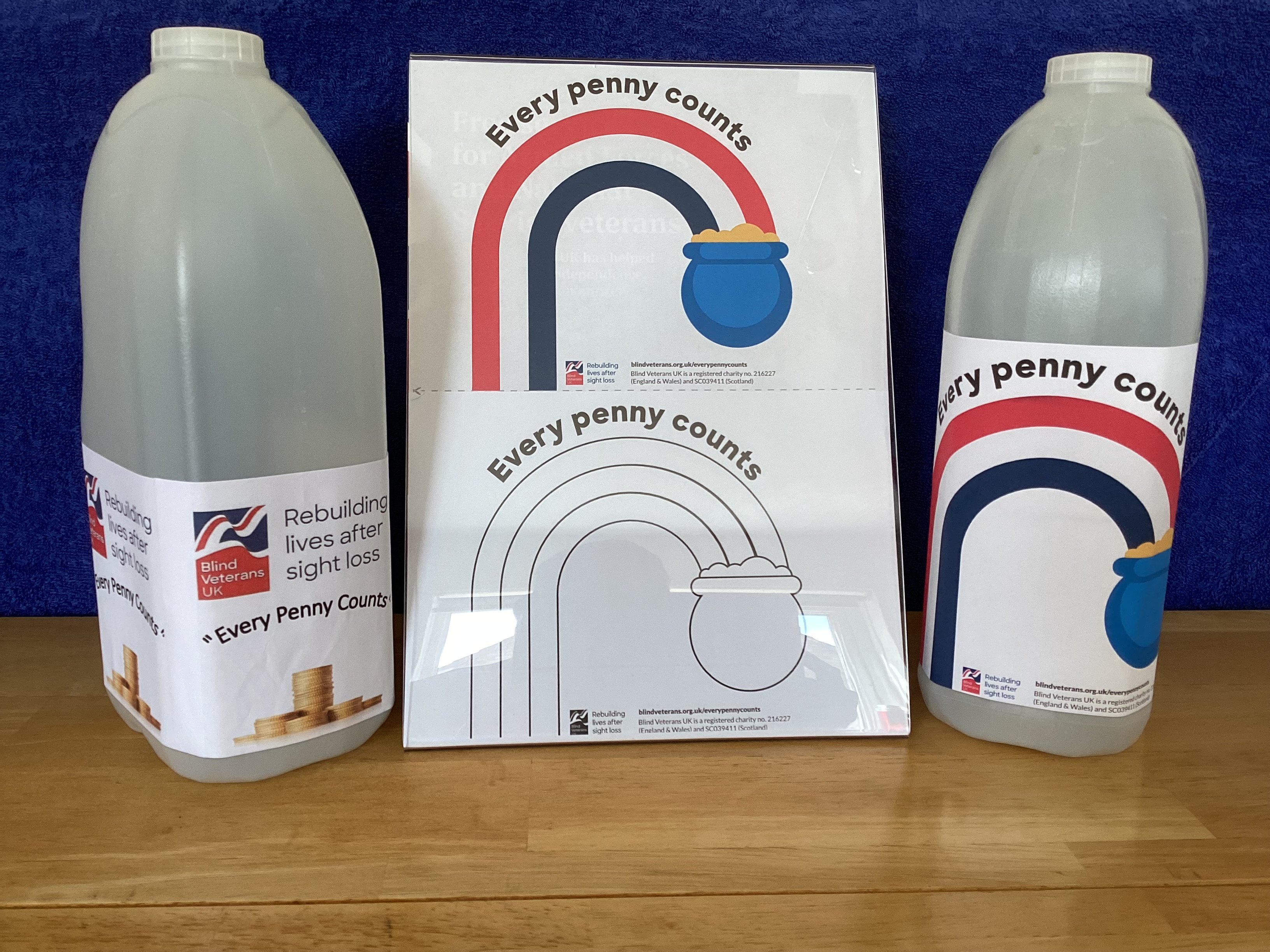 Two empty four pint plastic milk cartons displaying the 'Every Penny Counts' labels. One says 'Every Penny Counts' with a picture of coins stacked up and the other says 'Every Penny Counts' with a rainbow consisting of the Blind Veterans UK colours leading into a pot of gold.