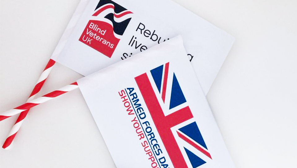 Two hand waving flags; one with the National Armed Forces Day logo lay across a second flag with Blind Veterans UK's logo on it. 