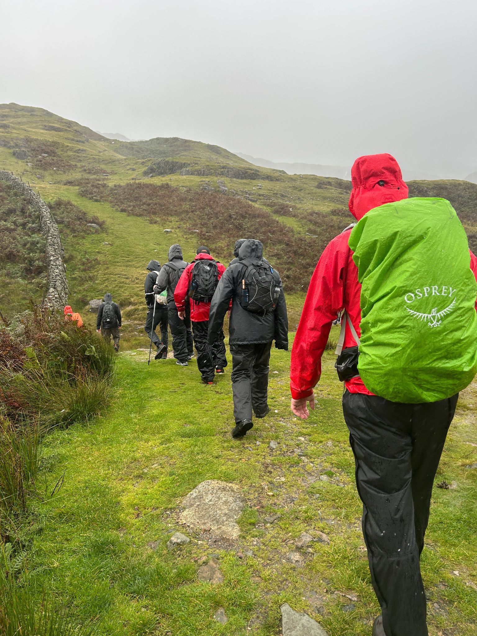 A picture taken from behind the group as they walk in single file across a hilly landscape. The rain is coming down and they all have their rain coats on and hoods up.