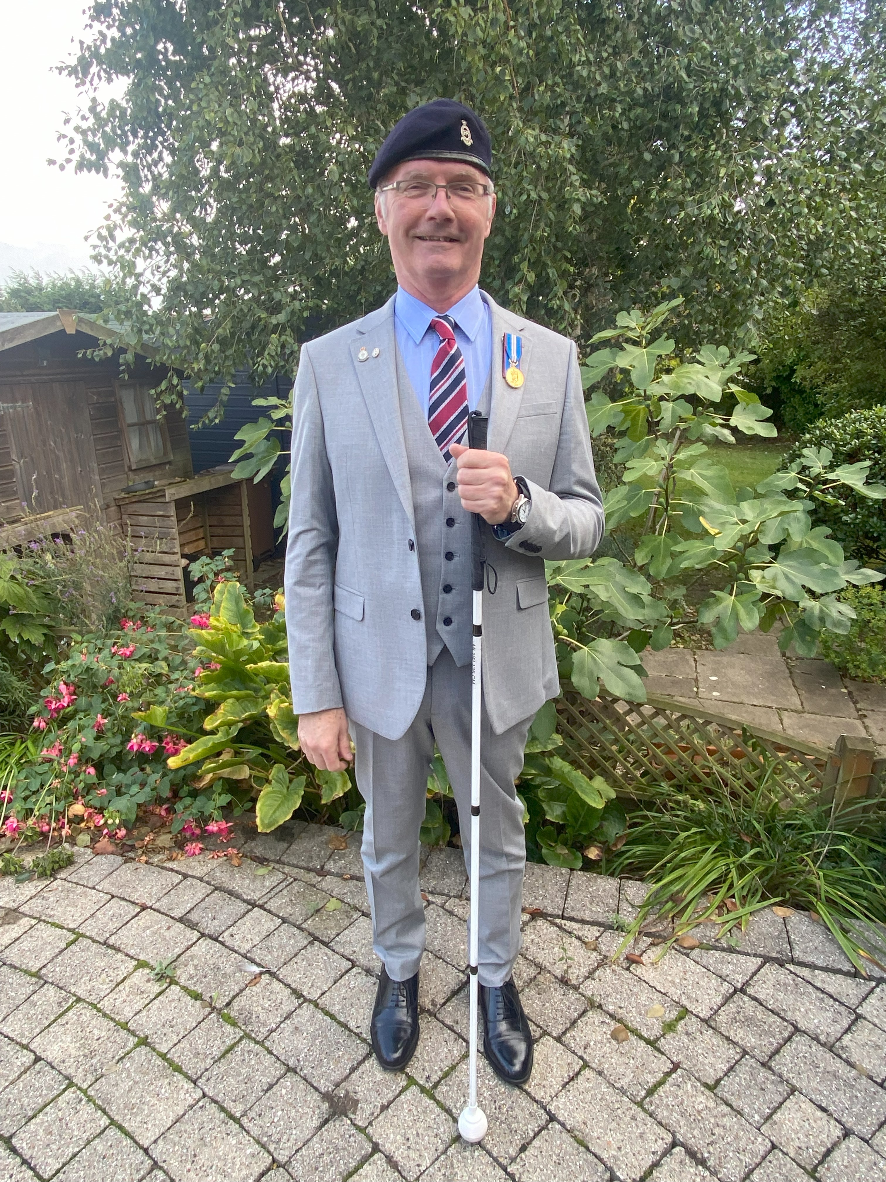 Mark stands in his garden wearing a grey suit, beret and medals. He is holding a white cane. 