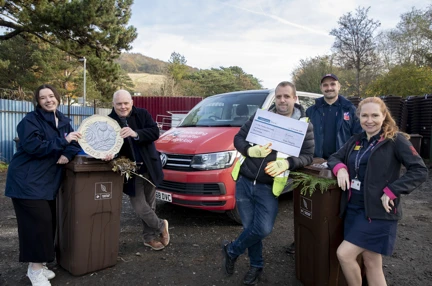 A picture of people leaning against two brown wheelie bins. One group are holding a cheque and the other a large £1 coin.