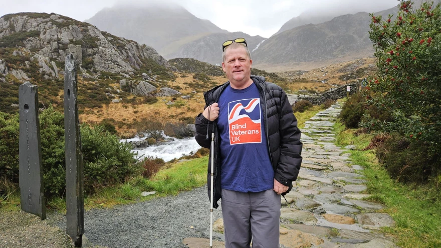 Phil on a cobble stone path with mountains behind him. He has yellow tinted glasses on his head and is holding his long cane. He is wearing a Blind Veterans UK T-shirt, grey walking trousers and a black jacket.