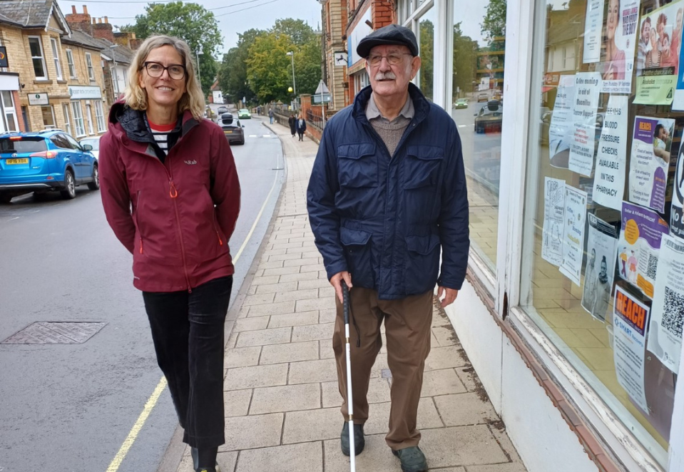 Blind Veteran Martin walking down a street, with a support worker. He is using a cane
