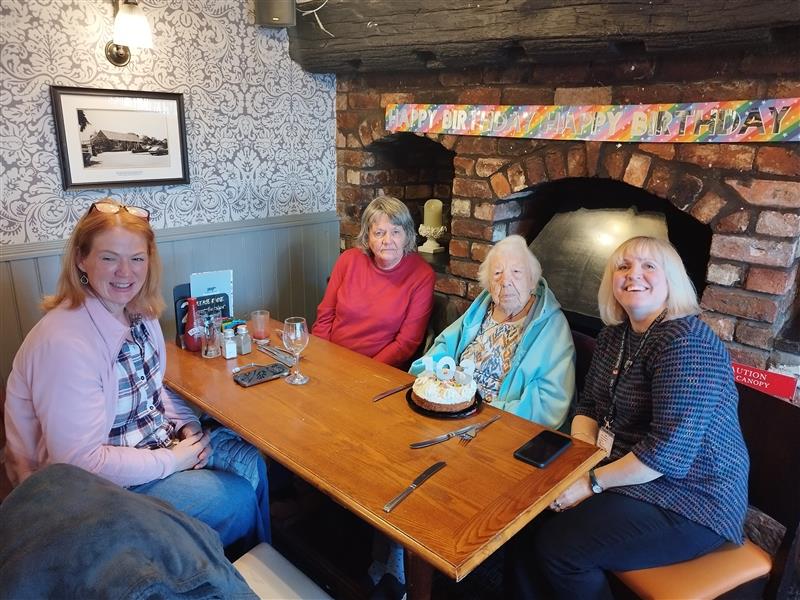 Olly seated at a restaurant table with volunteer Lanie, two friends and a birthday cake. They are all smiling in the cosy setting. A birthday banner is up behind them.