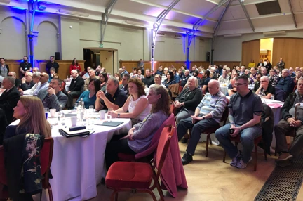 A photo of a group of blind veterans, family, firends and staff sitting round tables listening to a talk