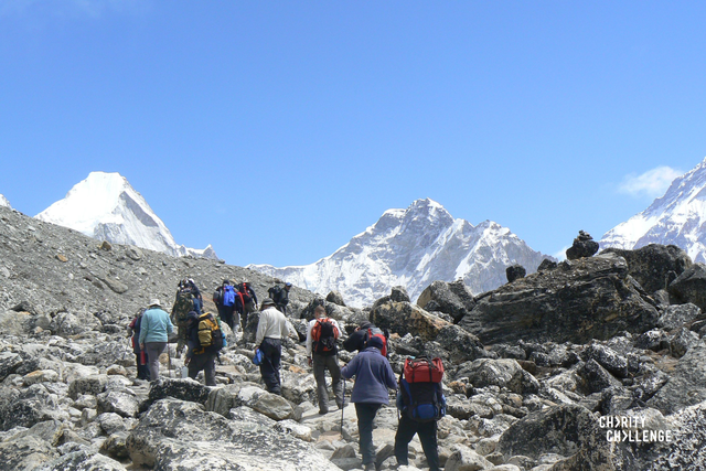 Group of people walking away from the camera across rocky snow covered landscape, heading towards towering, icy mountains that fill the background of the image.