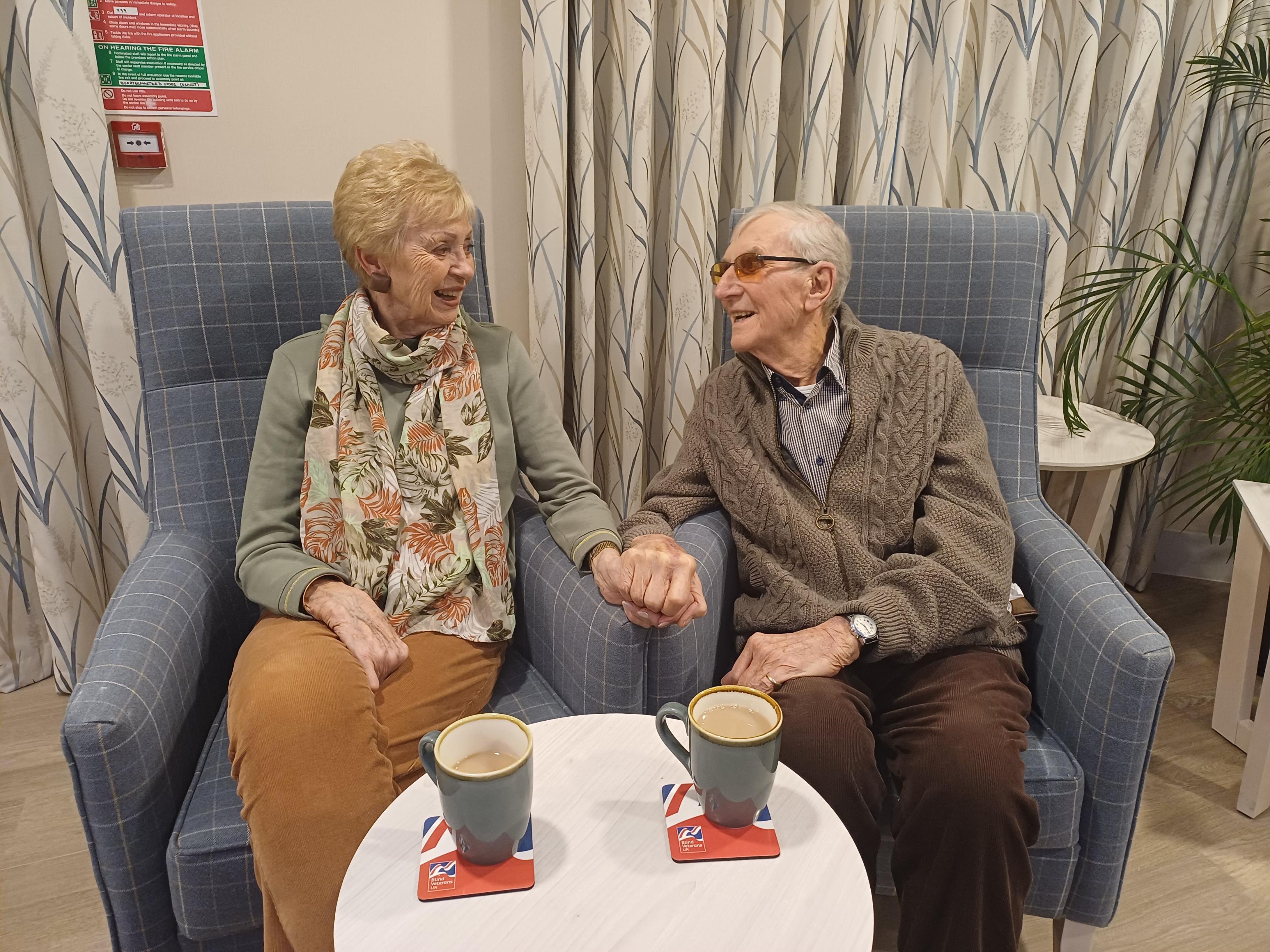 Blind veteran Jim and his wife Elsa sit on arm chairs next to each as they hold hands and look at one another with loving smiles