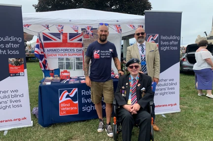 A Blind Veterans UK display stand set up with leaflets and posters with members of staff and blind veteran David stood in front