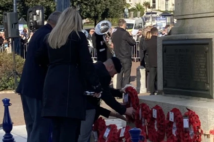 Billy dressed in smart clothes, bending over to lay his poppy wreath beside others. A military band is playing in the background.