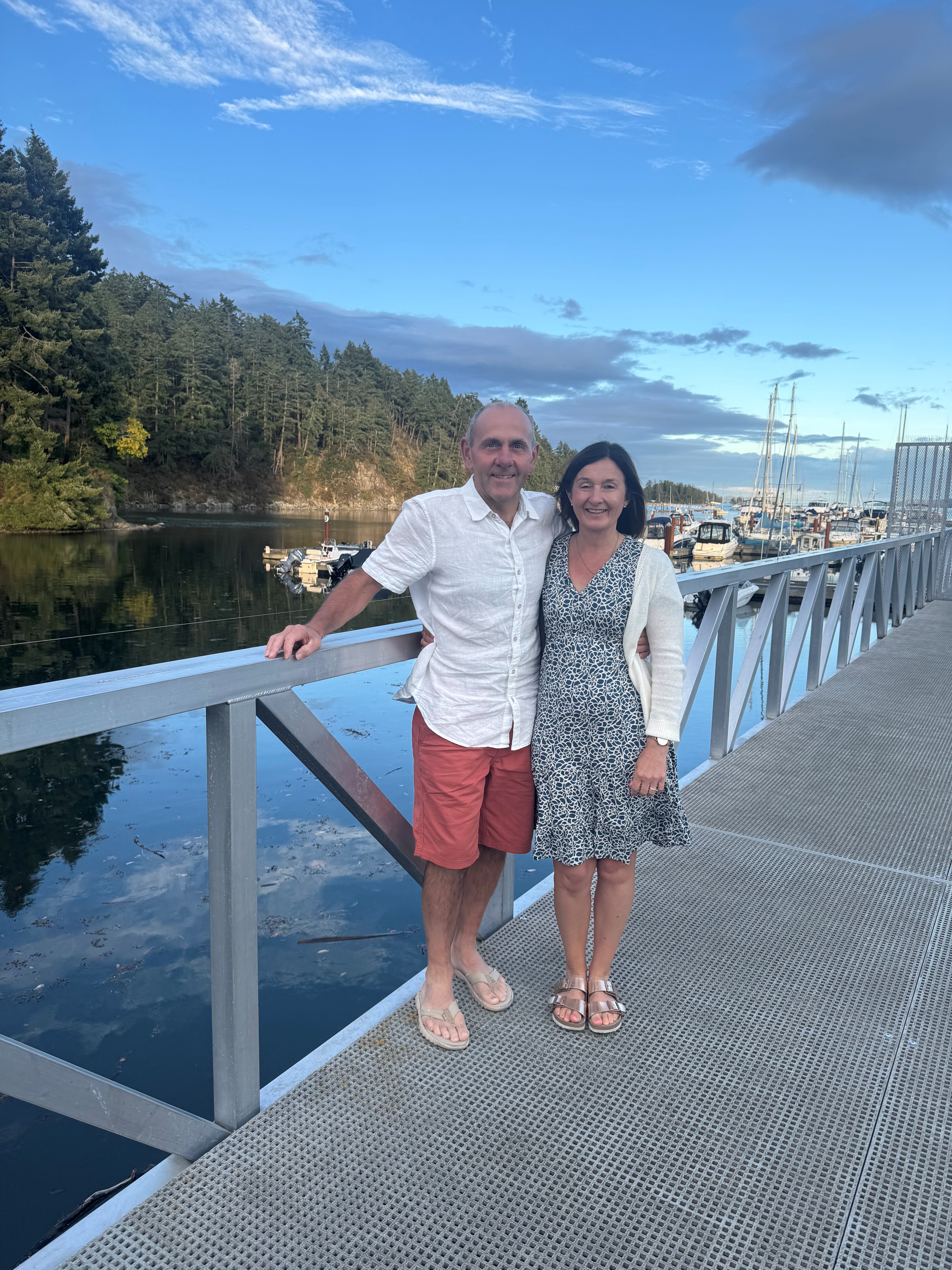 Kathryn and Simon are stood with their arms around one another in smart summer clothes next to a body of water with boats moored 