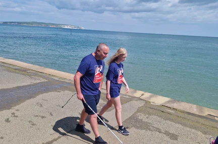 Ian wears a Blind Veterans UK t-shirt as he walks alongside his daughter. Ian is holding his white cane as they make their way along the promenade with the sea beside them.
