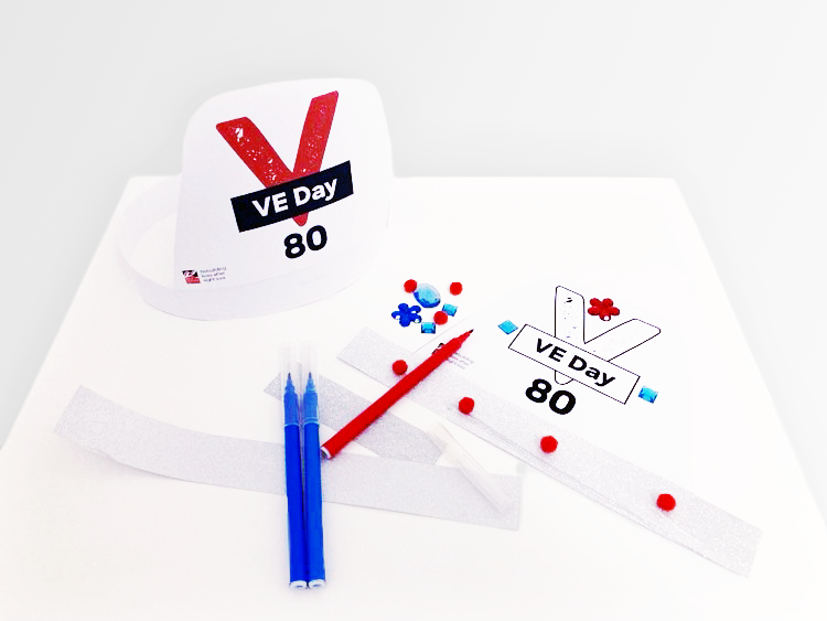 Assembled VE Day 80 party hat and one in the progress of being decorated with pom-poms, craft jewels and silver paper.