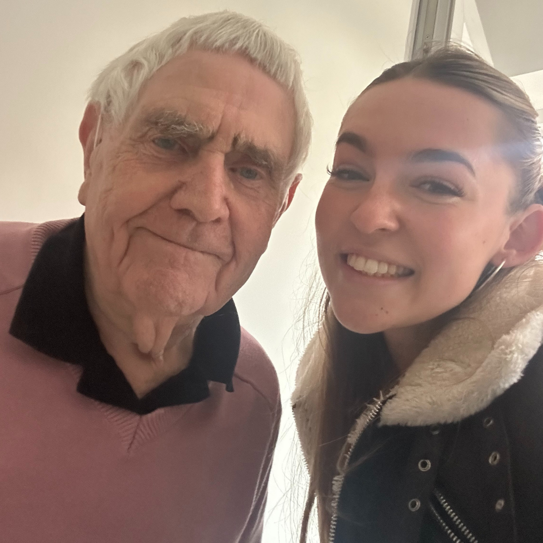 An elderly gentleman with grey hair stands next to a young lady with dark brown hair. They are looking at the camera and smiling.