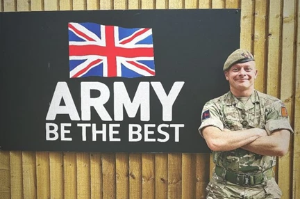 James in his Army uniform stood in front of a sign that says "Army - Be the Best" and a picture of a Union Jack flag.