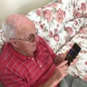 Blind veteran Ron, sitting on a sofa, holding a phone with synapptic software. The screen is displaying a large text main menu.