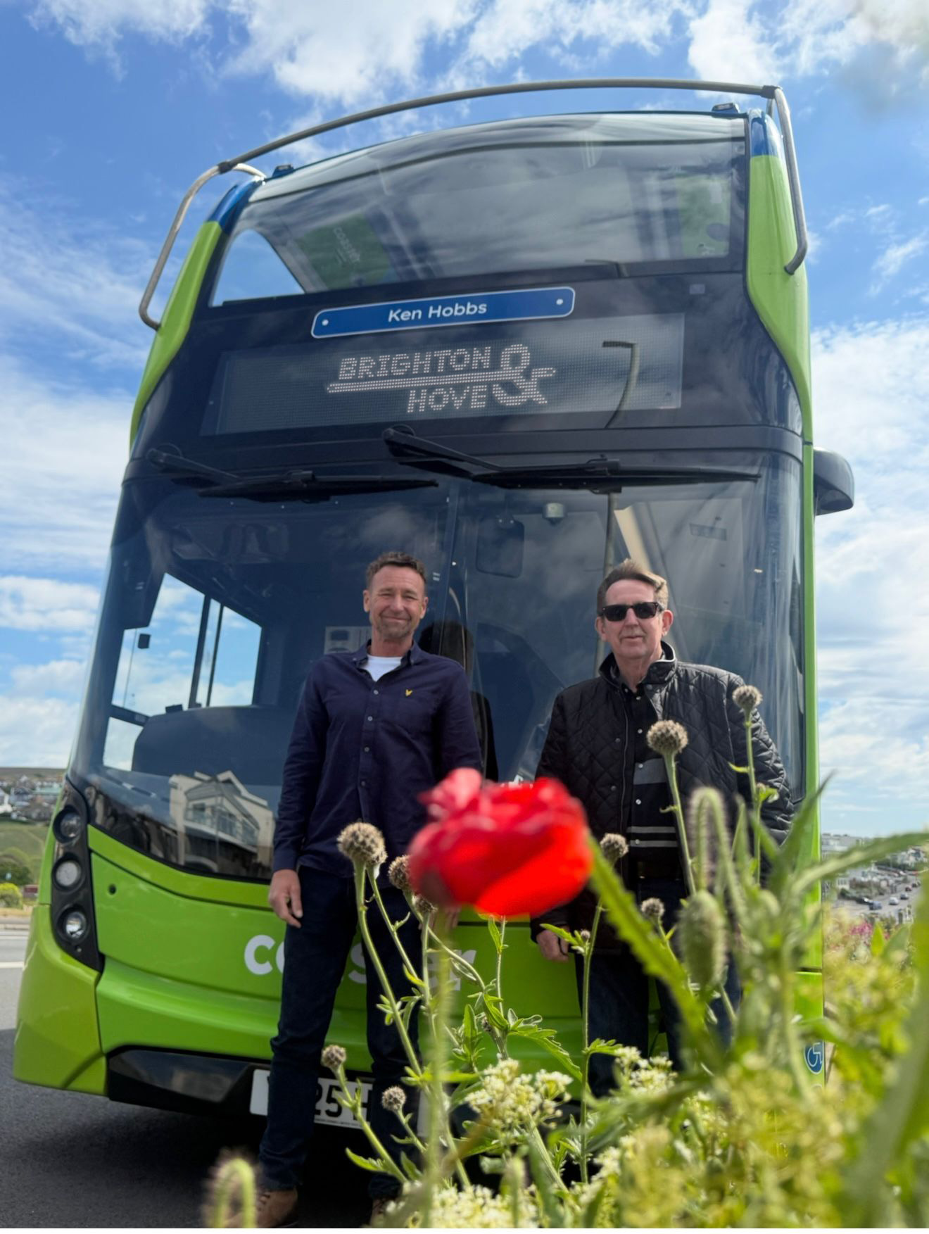 Two people standing in front of a Brighton & Hove Buses Coaster bus named after the late blind veteran Ken.