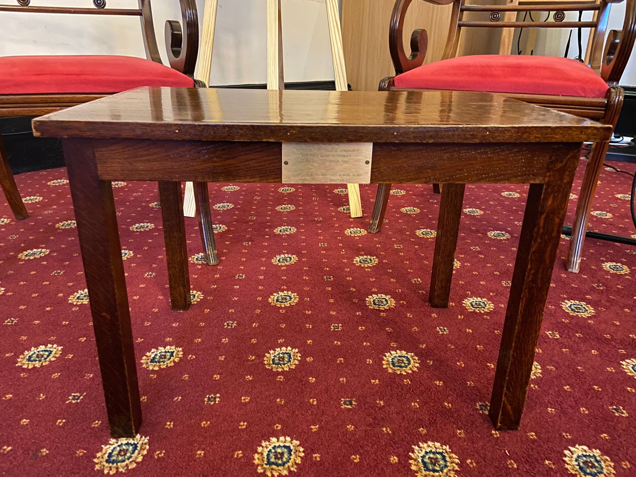  A small oak coffee table with a brass plaque on the front stands on a red patterned carpet, with red upholstered chairs behind inside a Sandhurst room.
