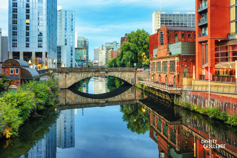 A stretch of Manchester Canal with colourful buildings either side and a footbridge stretching over the water.  The water is clear and still and the buildings, bridge and sky are reflected in the surface.