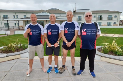 Four blind veterans wearing their Blind Veterans UK t shirts stood together in the garden with the Rustington Centre behind them.