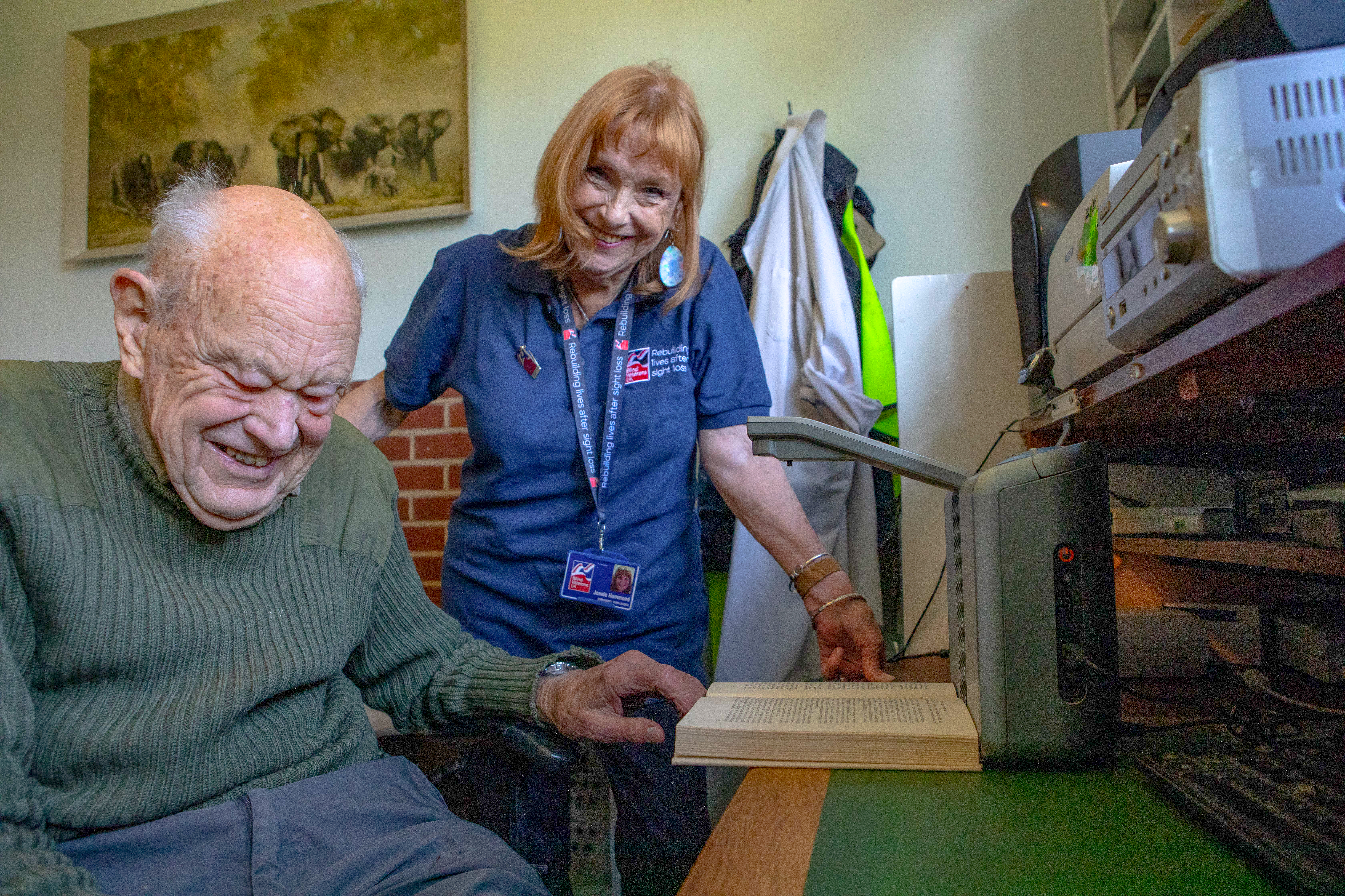 Jennie and Mike are both laughing and looking into camera as Mike puts a book under the reading device