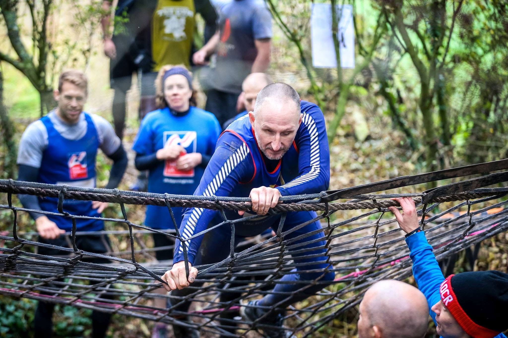 A person clambering across a horizontal rope net in the woods while others look on.