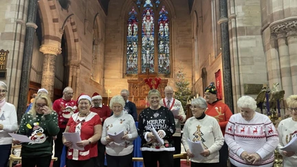 A group of people standing at the front of a church singing as part of a carol service, there is a stained glass window behind them
