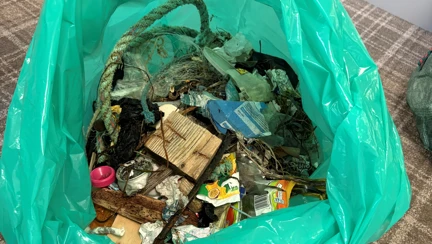 A green bin bag is open to show the assortment of litter collected from the beach it includes rope, fishing line, food packaging, small pieces of plastic and lots more unidentifiable pieces.