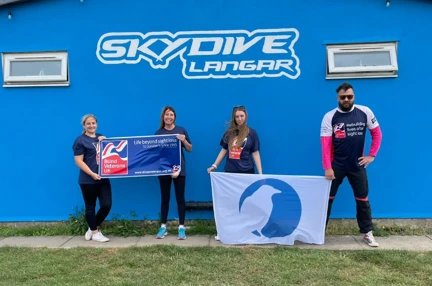Olivia stands with three of her colleagues, all wearing Blind Veterans UK t-shirts. They are under a sign that reads 'Skydive Langar'