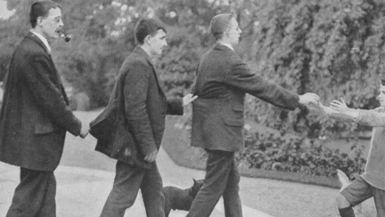 A historic black and white photograph of vision-impaired Belgian soldiers walking in a line, each holding onto the back of the man in front. A young boy at the front reaches out to guide them forward.