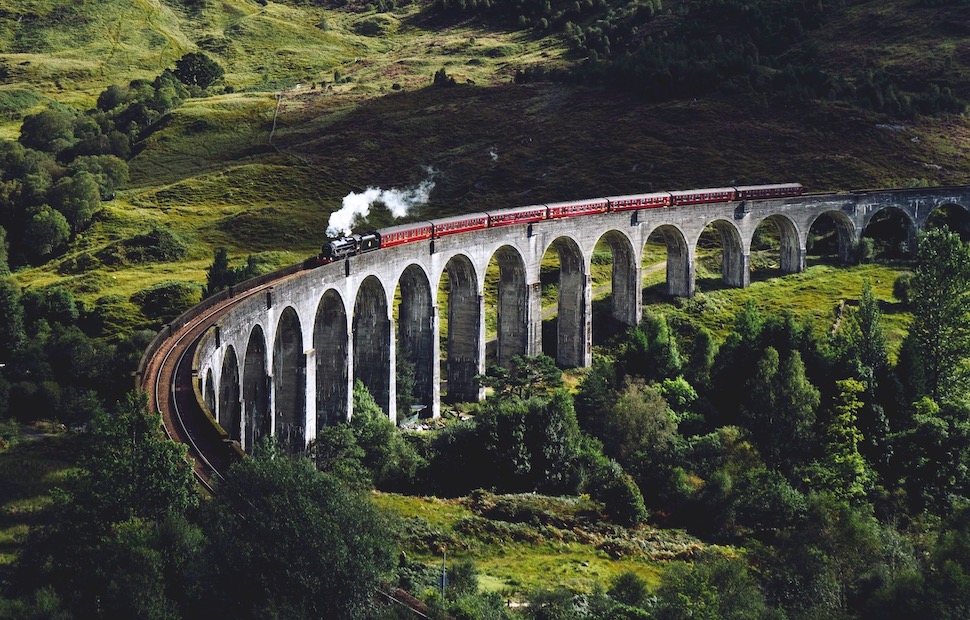 A steam train travelling over the Glenfinnan Viaduct, overlooking green fields and hills.