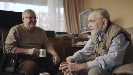 Volunteer Liam is holding a Blind Veterans UK mug as he smiles at blind veteran Ken, both are sitting down in Ken's living room