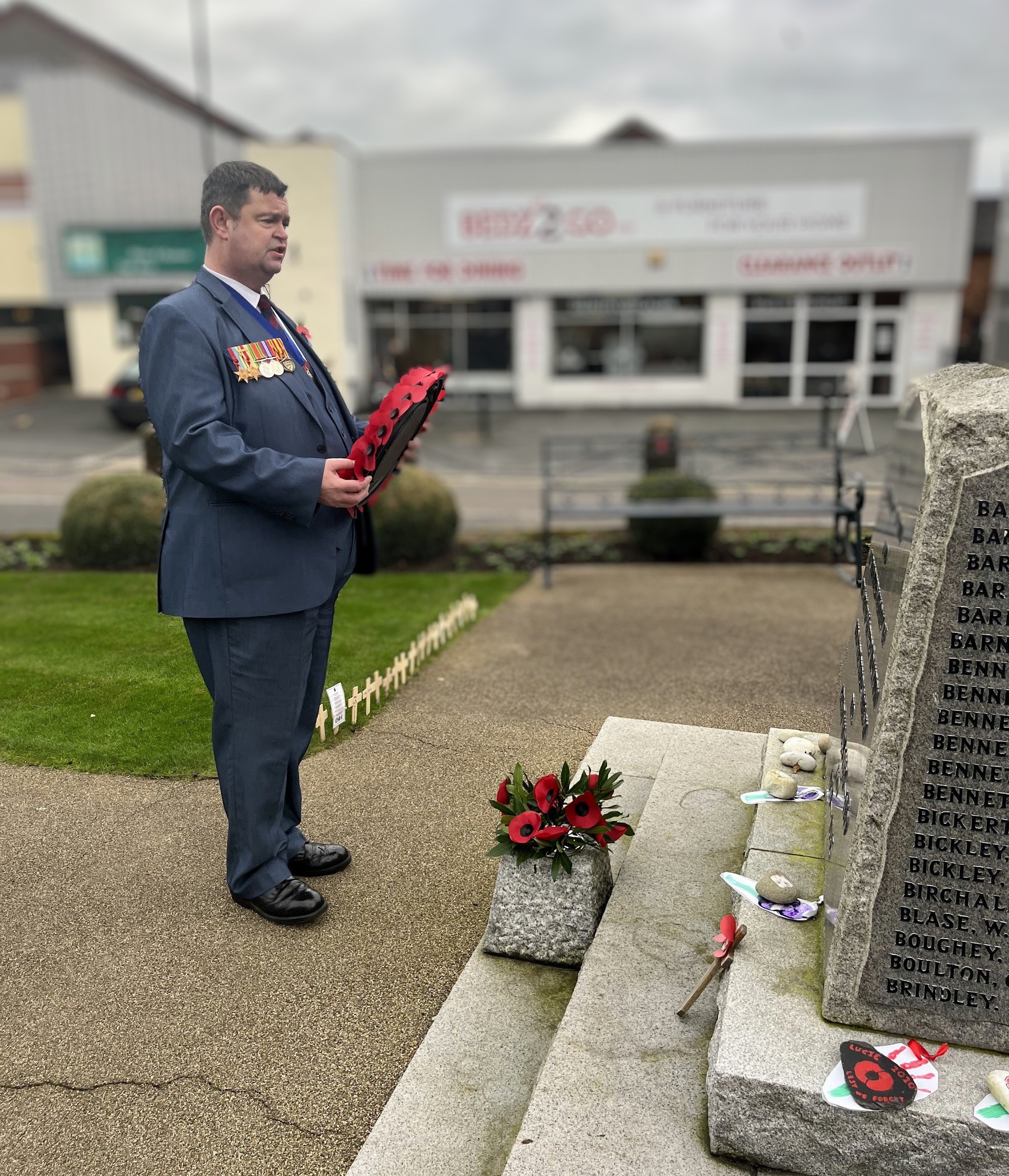 Clive stands in front of the war memorial and holds a wreath, ready to place it, he is wearing a blazer and medals