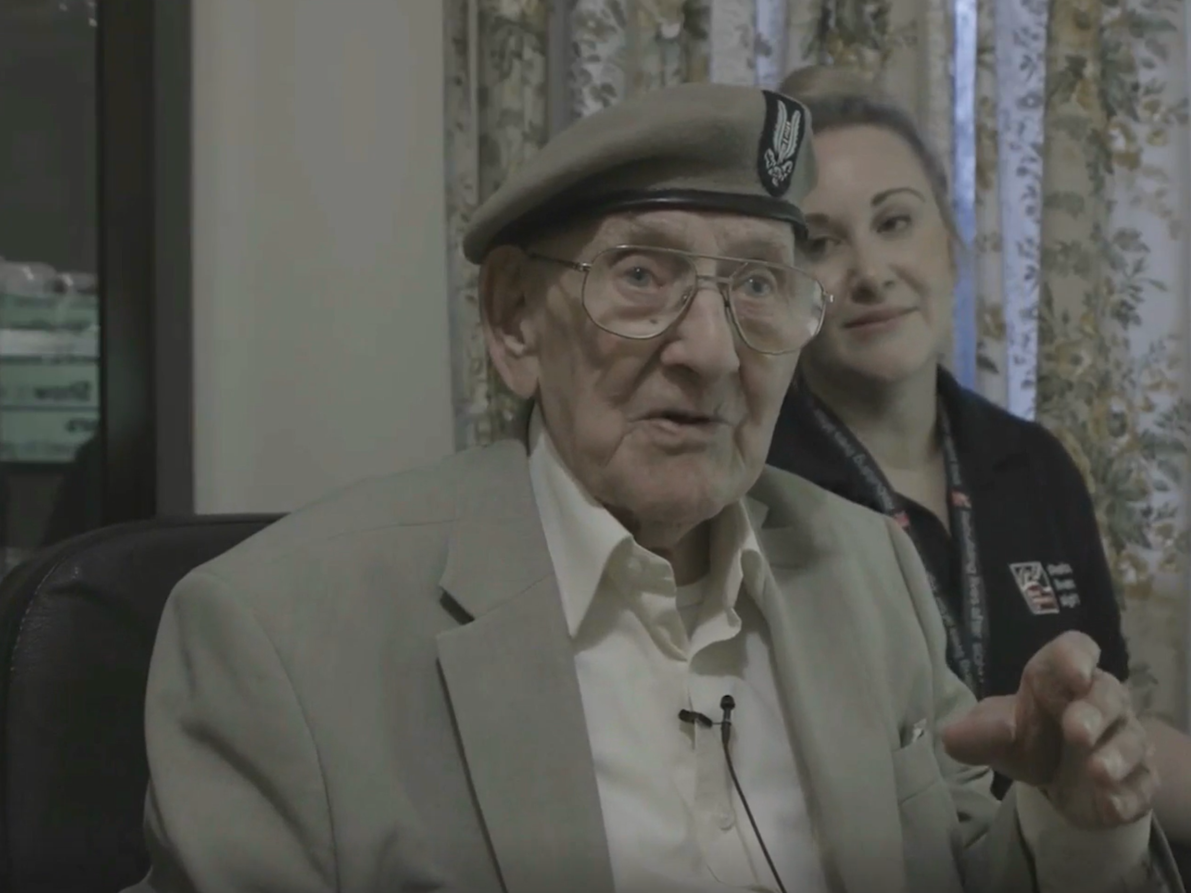 Blind veteran Matt sits at home, wearing a blazer and military beret, as he speaks about support from the charity. A Blind Veterans UK Community Support Worker sits next to him.