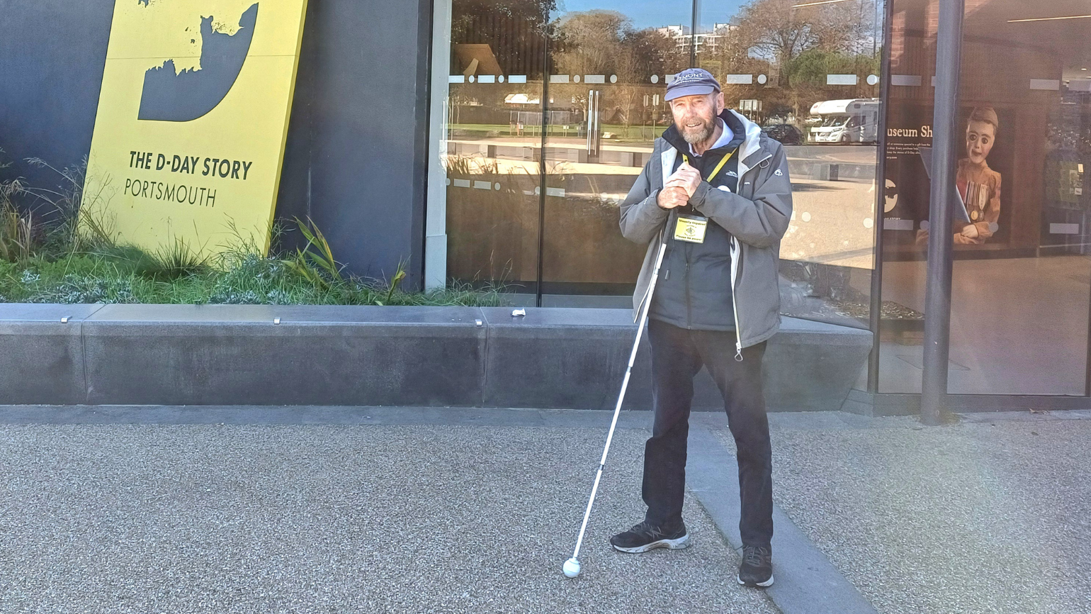 Dave stands smiling at the entrance of the D-Day museum, white cane in hand.