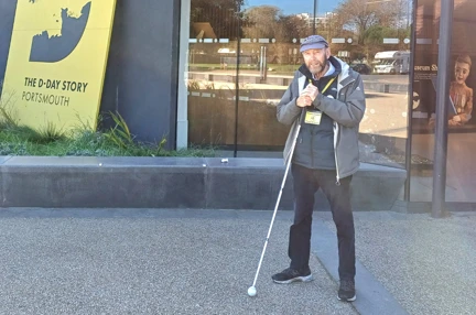 Dave stands smiling at the entrance of the D-Day museum, white cane in hand.