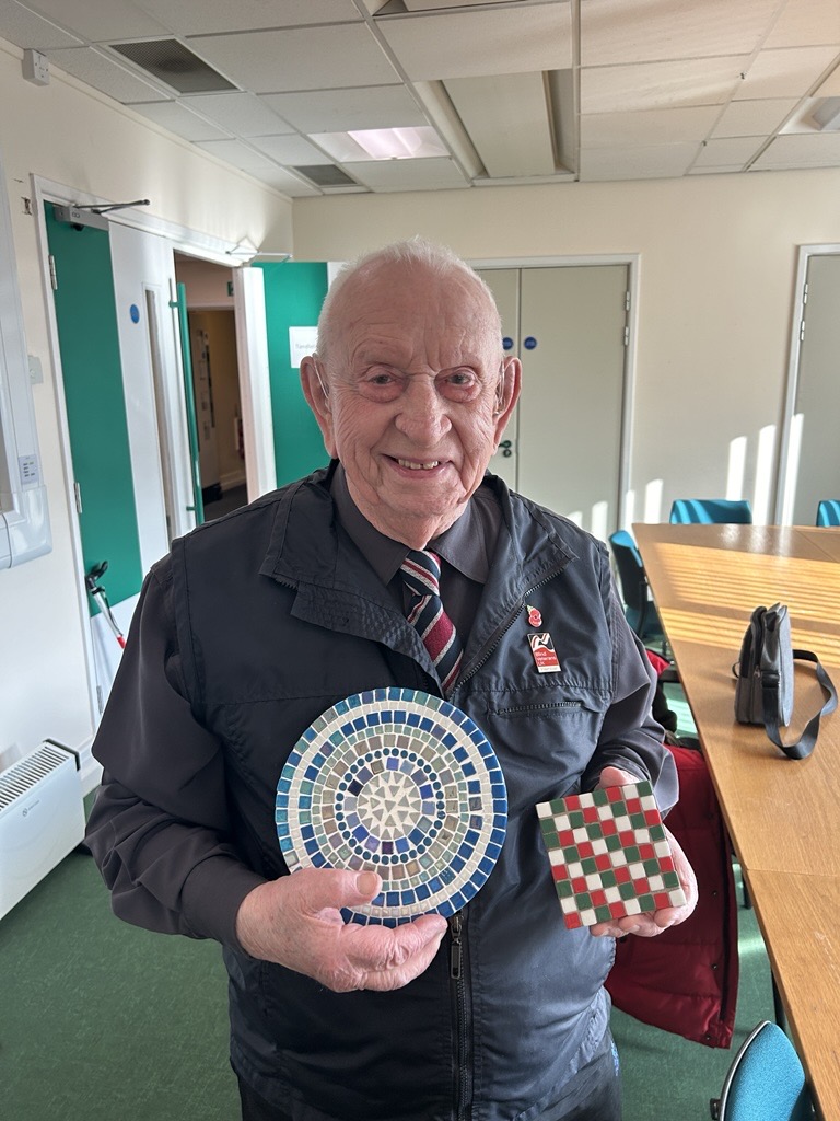 Glynne stands smiling while holding two mosaic projects he created