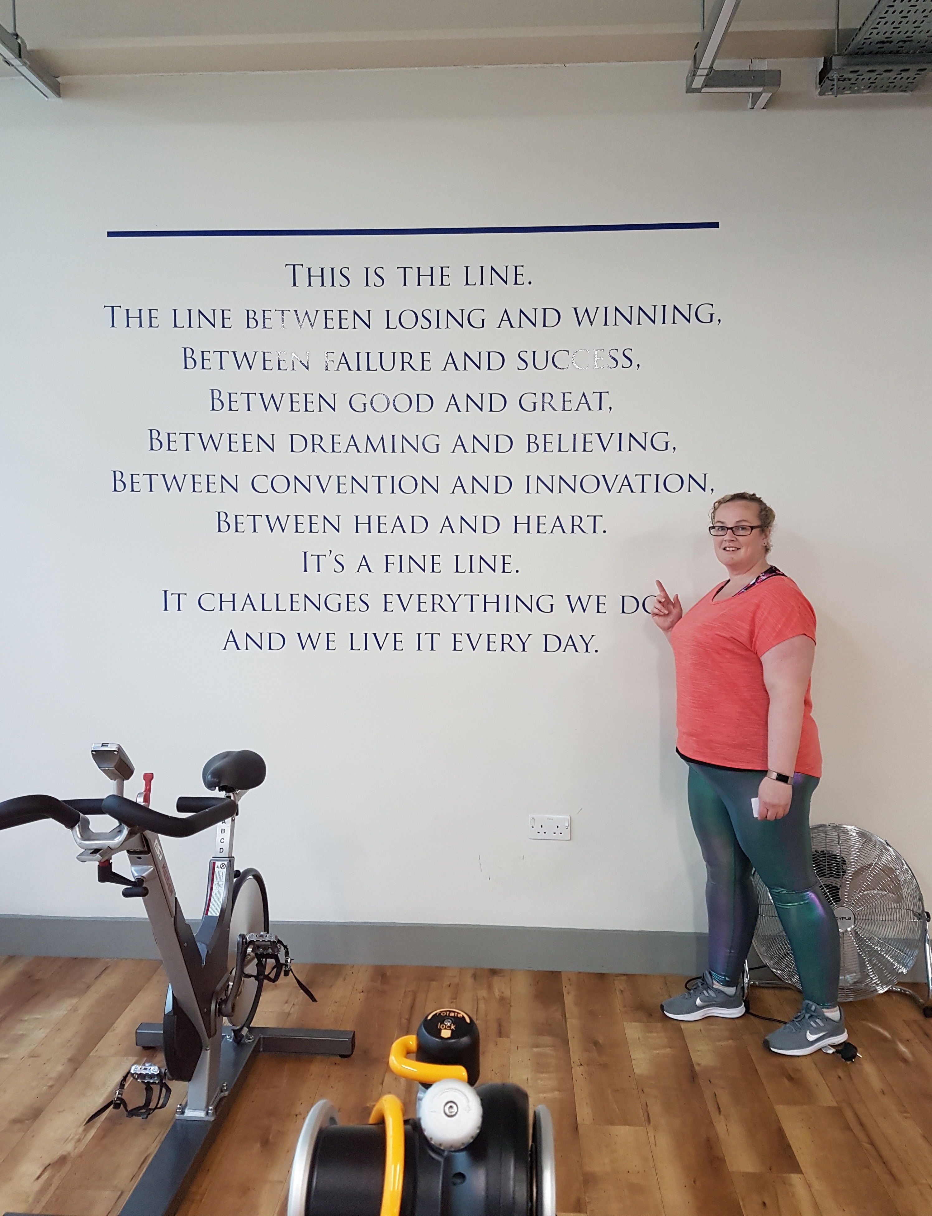 A photo of blind veteran Clare standing next to training equipment and pointing to inspirational message on wall