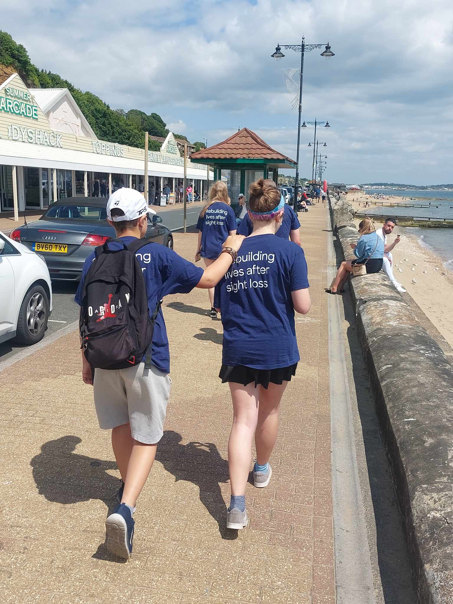 An image of the back of two walkers as they make their way along the seafront. A woman is acting as the guide and a man is holding on to her shoulder and walking slightly behind
