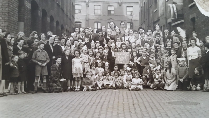 A black and white photograph showing a large group of women and children gathered in a cobbled street, smiling and wearing paper hats to celebrate VE Day. In the centre, a child holds a sign which reads: Mansford Street Party VE Day 1945. Blind veteran Thomas stands on the left, the only man in the photo, home on leave and in unfiorm.