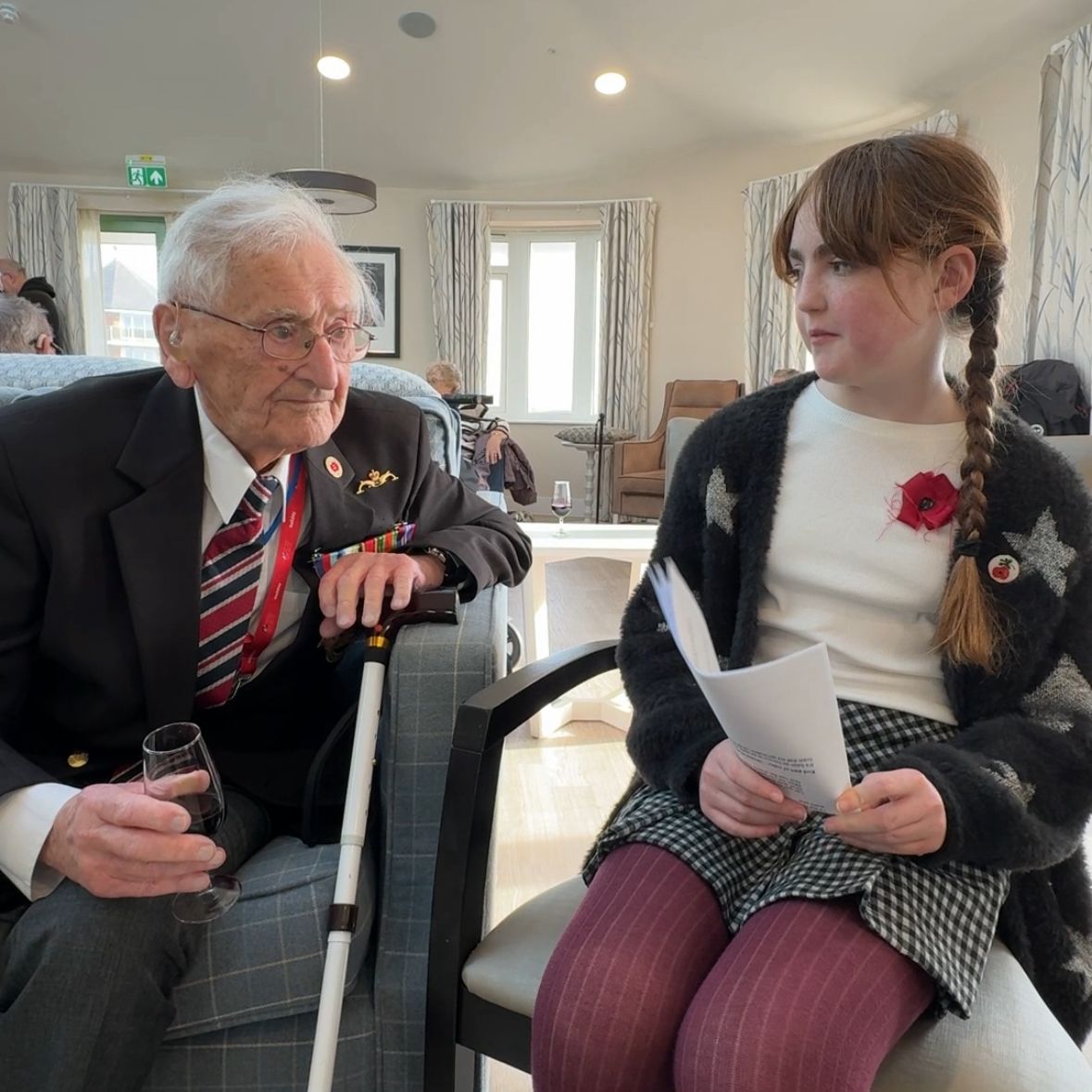 John and Evelyn sit in chairs next to each other having a discussion. Evelyn is wearing a poppy and John is wearing his medals