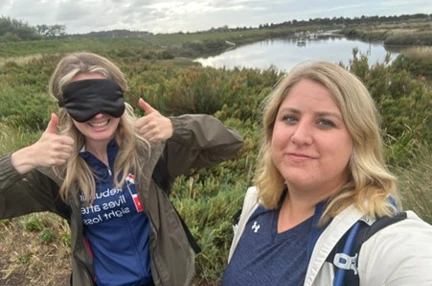 Rosie is wearing a blindfold and has two thumbs up and a huge smile. Kat is standing next to her and behind them there are green fields and a lake.
