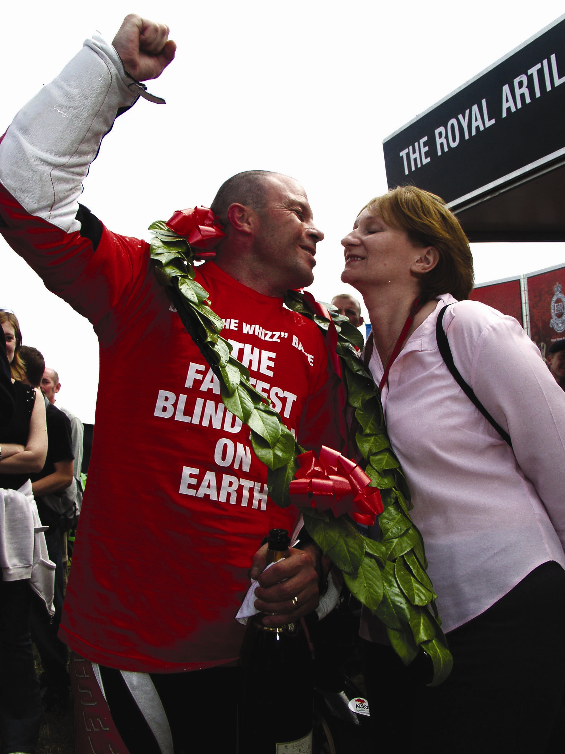 Blind veteran Billy, cheering in celebration while wearing motorcycle gear and holding a bottle of champagne as he leans in to kiss his wife