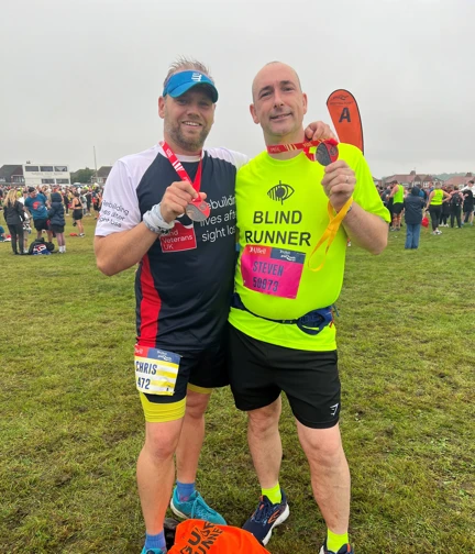 Blind veteran Steve and his guide runner Chris are holding up their medals to the camera. They are stood with their arms around one another's shoulders. Chris is wearing a Blind Veterans UK running top and Steve is wearing a bright yellow t-shirt with "blind runner" written on it.