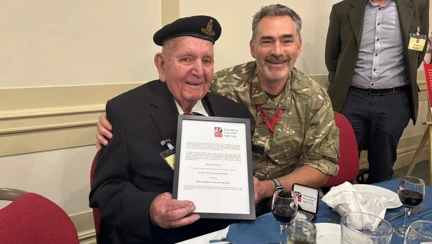 A WWII veteran proudly holding a certificate, seated beside a man in military dress.